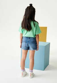 Camiseta corta verde de manga corta, pantalones cortos de mezclilla cortados, zapatillas blancas con acentos en tonos pastel. Dos bloques geométricos en el fondo.