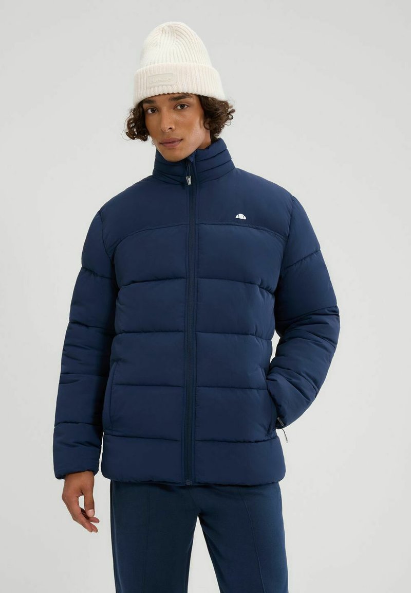 Chaqueta acolchada en azul marino con cuello alto, diseño acolchado y cremallera en la parte delantera. Llevada con un gorro de punto color crema para mayor calidez.