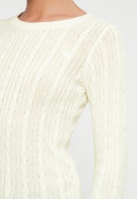 Close-up van een persoon die een witte kabelgebreide trui draagt met verticale vlechtpatronen en een ronde hals.