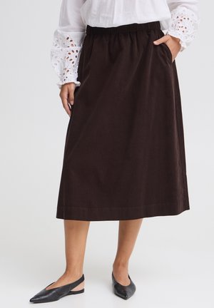 Femme portant une jupe midi marron foncé avec taille élastique, un chemisier blanc à manches ajourées et des ballerines slingback noires, debout les mains dans les poches.