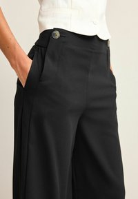 Pantalon noir à jambes larges en tissu lisse, doté de poches latérales et de deux boutons décoratifs à la taille. Coupe ajustée, design minimaliste.