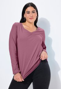 Halenka s dlouhým rukávem a výstřihem do V z jemné mauve tkaniny; má volný střih a boční rozparky, kombinovaná s tmavými džíny. Žádné viditelné vzory ani kovové detaily.