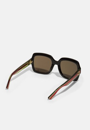 Gafas de sol grandes y negras con lentes marrones. Cuenta con brazos a rayas únicos en verde y rojo, y acentos en dorado. Forma cuadrada.