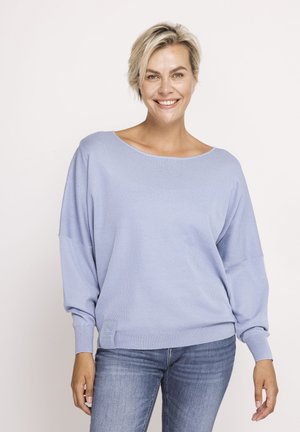 Lächelnde Frau mit kurzen blonden Haaren, die einen locker sitzenden hellvioletten Pullover und blaue Jeans trägt, steht vor einem schlichten weißen Hintergrund.