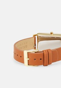 Hnědý kožený náramkový hodinky s obdélníkovým zlaceným pouzdrem. Texturovaný náramek s přezkovým zapínáním, minimalistiký ciferník s průhledným sklem.