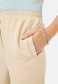 Beige jogger pants with an elastic waistband, side pockets, and a smooth texture. A hand rests in the pocket, wearing a silver beaded bracelet.