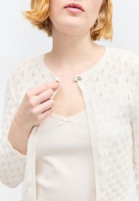 Femme aux cheveux blonds courts boutonnant le bouton supérieur d'un cardigan blanc texturé par-dessus un débardeur blanc.