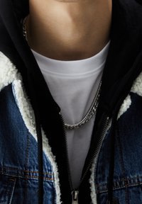 Primer plano de una persona que lleva una camiseta blanca, un collar de cadena plateada, una sudadera con capucha negra y una chaqueta de mezclilla azul con forro de lana blanca.