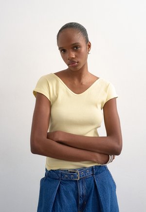 Γυναίκα με σκουρόχρωμο δέρμα και μαλλιά τραβηγμένα προς τα πίσω, φοράει ανοιχτό κίτρινο κοντομάνικο μπλουζάκι και μπλε ψηλόμεσα τζιν με ζώνη, σταυρωμένα χέρια.