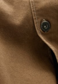 Brown fabric with a fine ribbed texture, featuring white stitching and a round metal snap closure. The surface exhibits a subtle sheen.