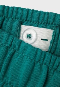 Tealfärgad stickad midjeband med samlad struktur, med en vit tygetikett och en genomskinlig knapp med en grön tråddekor i mitten.