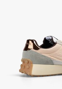 Parte trasera de una zapatilla beige y gris con lengüeta de talón metálica en oro rosa y suela de goma texturizada con tachuelas hexagonales sobre fondo blanco.