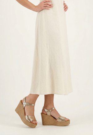 Vrouw draagt beige sleehak sandalen met enkelriem en een gestructureerd plateau, en een crèmekleurige midi-rok, staand tegen een effen achtergrond.