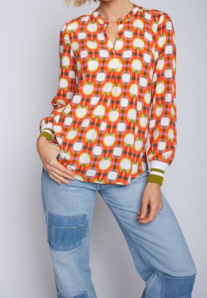 Žena v barevné blůze s geometrickým vzorem v oranžové, bílé a zelené barvě, doplněná světle modrými džínami s riflovými záplatami.