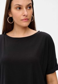 Czarna, oversize koszulka z okrągłym dekoltem i podwiniętymi krótkimi rękawami. Gładki materiał z delikatnym połyskiem i luźny krój. Okrągłe srebrne kolczyki.