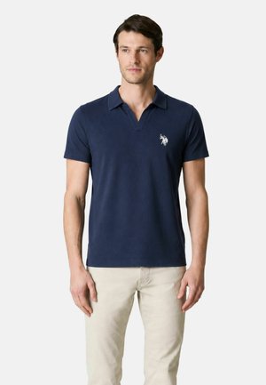 Uomo che indossa una polo blu navy a maniche corte con un logo bianco e pantaloni beige, in piedi davanti a uno sfondo chiaro e uniforme.