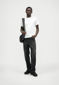 T-shirt branca com o logotipo "REPLAY", jeans de ganga pretos e ténis pretos. Modelo segura uma mala transversal preta, de pé contra um fundo neutro.