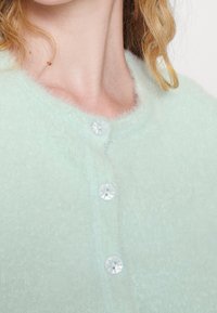 Mátově zelený, nadýchaný cardigan s kulatým výstřihem, zdobený průhlednými plastovými knoflíky a měkkou texturou. Detailní záběr horní části.