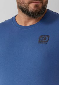 Niebieski bawełnowy T-shirt z okrągłym dekoltem, z czarnym logo przedstawiającym glob i napisem "Discovery" na górnej lewej części klatki piersiowej.