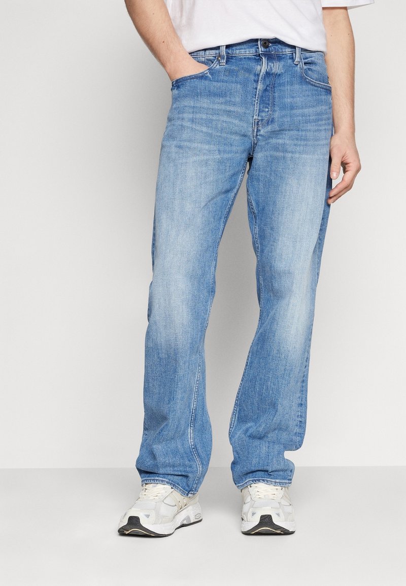Hombre vistiendo jeans rectos de color azul claro y zapatillas blancas, de pie con una mano en el bolsillo frente a un fondo liso.