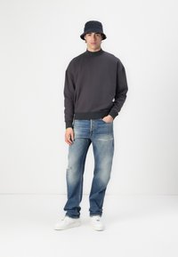 Mørkegrå sweatshirt med høj krave, kombineret med lyseblå slidte jeans og hvide sneakers. Model bærer en sort bøttehat.