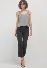Femme aux cheveux bouclés portant un haut en tricot sans manches gris, un jean cropped noir taille haute et des escarpins noirs à brides de cheville, marchant en avant.