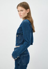 Chaqueta de mezclilla recortada en denim azul oscuro con mangas largas, un bolso en el pecho y acentos en botones dorados. Textura suave y diseño ajustado.
