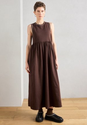 Abito lungo marrone senza maniche con gonna arricciata e impunture a contrasto lungo la scollatura. Indossato con zoccoli neri e calzini marroni.