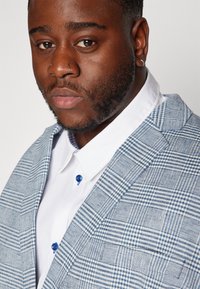 Μπλε και λευκή καρό σακάκι με υφή, συνοδευόμενο από ένα αντίθετο λευκό πουκάμισο και μπλε κουμπιά. Κοντινό πλάνο από τον γιακά και το πέτο.