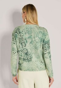 Vrouw met lang blond haar, gekleed in een groene trui met bloemenpatroon en crèmekleurige broek, gezien vanaf de achterkant tegen een neutrale achtergrond.