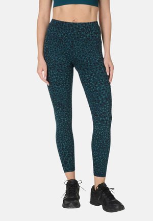 Vrouw draagt hooggetailleerde, donker teal leggings met zwart luipaardprintpatroon en zwarte sportschoenen, staand tegen een witte achtergrond.