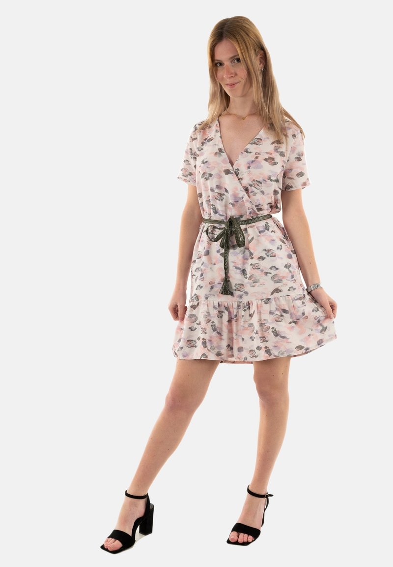 Vestido envolvente floral en rosa claro con detalles en verde, mangas cortas, dobladillo con volantes y cinturón a juego. Llevado con tacones negros de punta abierta.