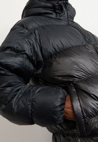 Black puffer jacket with a glossy finish, featuring a high collar, zip closure, and wide, quilted sections for insulation.