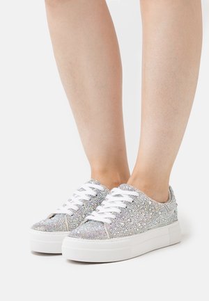 Jambes portant des baskets basses blanches à plateforme, ornées de petits strass argentés réfléchissants avec des lacets blancs, sur un fond clair uni.