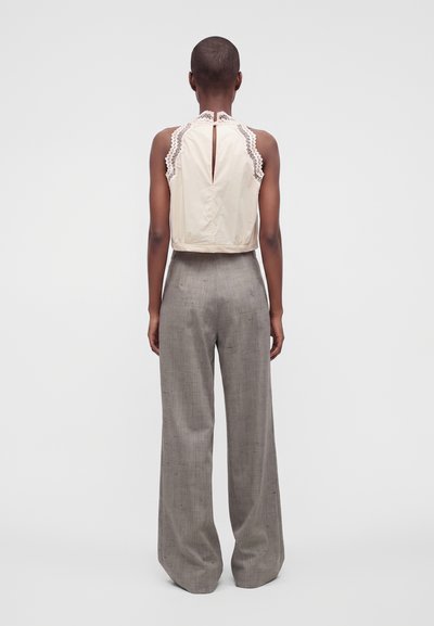 Personne portant un haut beige sans manches avec bordure en dentelle et détail en forme de trou dans le dos, associée à un pantalon large gris taille haute, debout de dos.
