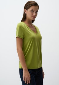 Zielona koszulka z krótkim rękawem w serek, wykonana z miękkiego materiału, o luźnym kroju i gładkiej fakturze, w połączeniu z ciemnoniebieskimi dżinsami.