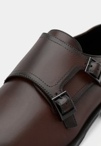 Dunkelbrauner Lederschuh mit doppelten Monk-Riemen und schwarzen Metallverschlüssen vor weißem Hintergrund.