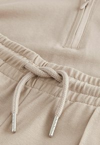Beige Jogginghose aus strukturiertem Stoff, mit elastischem Bund und einer geflochtenen Kordel mit silbernen Endstücken. Das Design umfasst eine Reißverschlusstasche.
