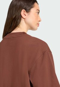 Camiseta de algodón marrón con un corte relajado, mangas cortas y un escote redondo. Textura suave, con un detalle mínimo, vista desde atrás.