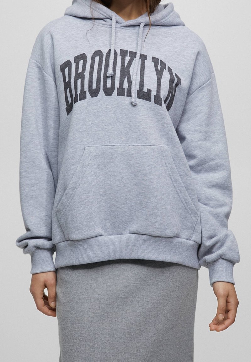 Personne portant un sweat à capuche gris clair avec le texte "BROOKLYN" en gris foncé, associé à une jupe gris clair assortie, debout devant un fond uni.