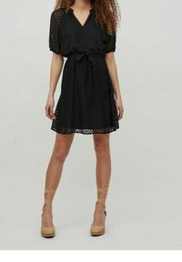 Zwarte jurk met een V-hals, korte pofmouwen en een strik taille. Getextureerde stof met een subtiel patroon. Gecombineerd met tan espadrille wedges.