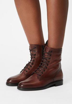 Botas de cuero marrón con cordones, de tobillo, con una textura suave, punta redonda y cremallera lateral. Presentan cordones resistentes y un tacón bajo.