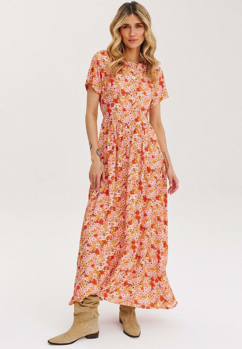 Kvetinové maxi šaty v ružovej, oranžovej a červenej farbe. Krátke rukávy, okrúhly výstrih, zúžený pás, splývavá sukňa, spárované s béžovými čižmami po kolená.