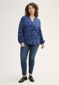 Blaue gemusterte Bluse mit V-Ausschnitt, langen Ärmeln und lockerer Passform, kombiniert mit dunklen Denim-Hosen und schwarzen Schnürschuhen.