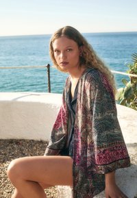 Zwiewny narzutka w stylu kimono o wielokolorowym wzorze paisley, wykonana z lekkiego materiału, z szerokimi rękawami i o długości do połowy uda.