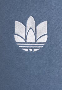 Blått tyg med en jämn textur pryds av en vit broderad logotyp som liknar en stiliserad blomma med tre kronblad och tre horisontella ränder.