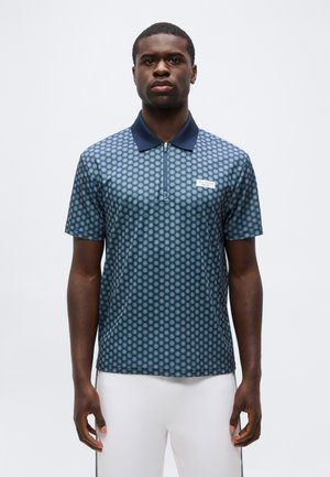 Mężczyzna ubrany w niebieskie, wzorzyste polo z krótkim rękawem, granatowym kołnierzem i białe spodnie, stoi na jednolitym jasnym tle.