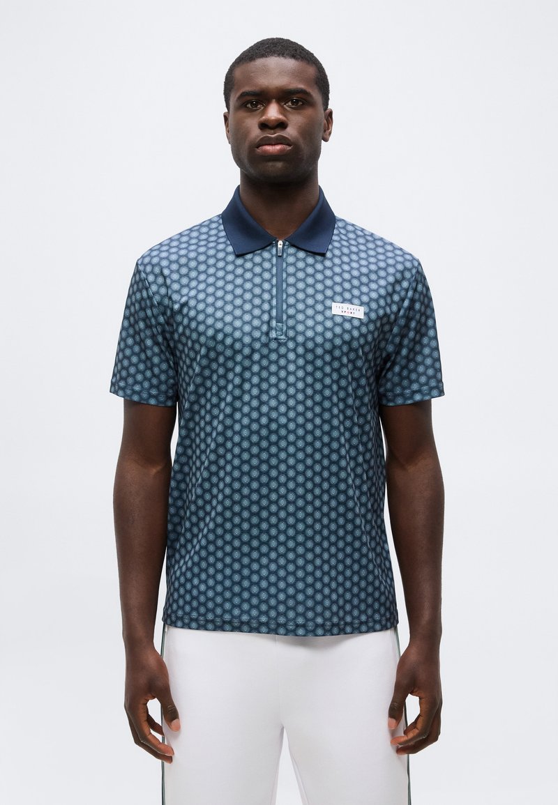 Άνδρας που φοράει μπλε μπλούζα πόλο με σύντομο μανίκι με σχέδιο, με σκούρο μπλε γιακά και άσπρο παντελόνι, στέκεται μπροστά σε απλό φωτεινό φόντο.