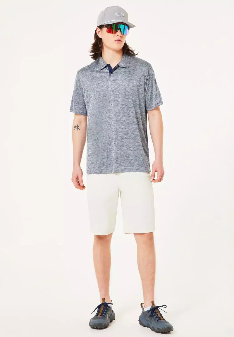 Hombre con polo gris, pantalones cortos blancos, gorra gris, gafas de sol deportivas de colores y zapatillas deportivas gris oscuro, de pie frente a un fondo liso.