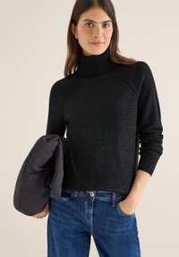 Schwarzer Rollkragenpullover mit gerippter Textur, Raglanärmeln und lockerer Passform. Blaue Jeans und eine graue Daunenjacke sind sichtbar.